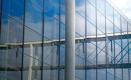 Großes, verglastes Gebäude, in dem sich blauer Himmel und Wolken spiegeln
