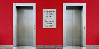 Eine rote Wand mit 2 Aufzugstüren und einer weißen HInweistafel zwischen den Aufzügen

