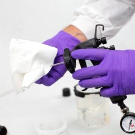 Hände mit violetten Handschuhen reinigen eine Lackierpistolennadel mit einem weißen Reinigungstuch